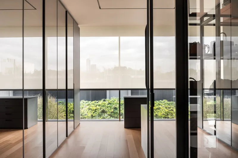 Glass-partitioned hallway with wooden flooring overlooking a terrace planted with low shrubs under hazy sky