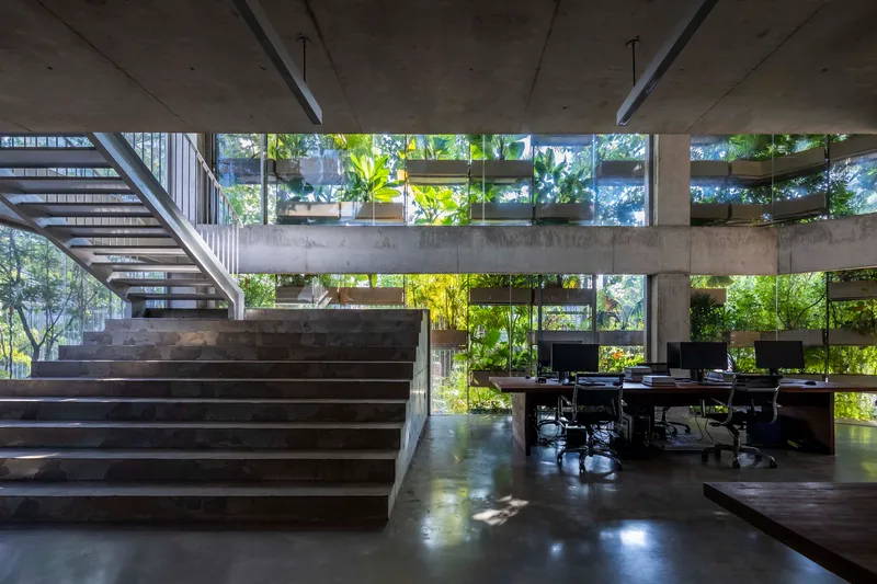Concrete staircase rising beside open workspace with desks below planted balcony edges visible through glazing