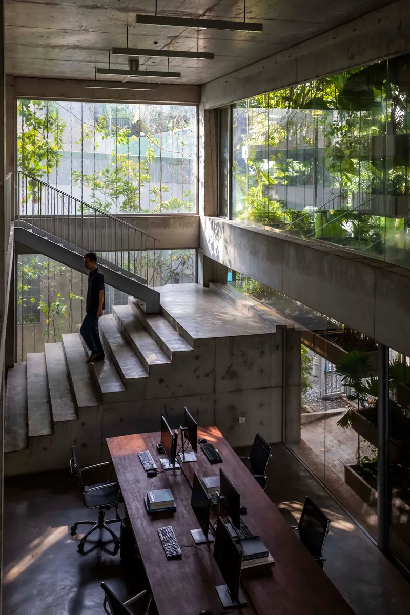 Concrete staircase ascending through multi-level workspace with person walking and vegetation outside windows