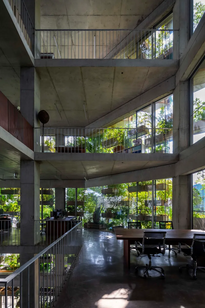 Interior view of stacked concrete floors with vertical rod railings and greenery visible through perimeter glass