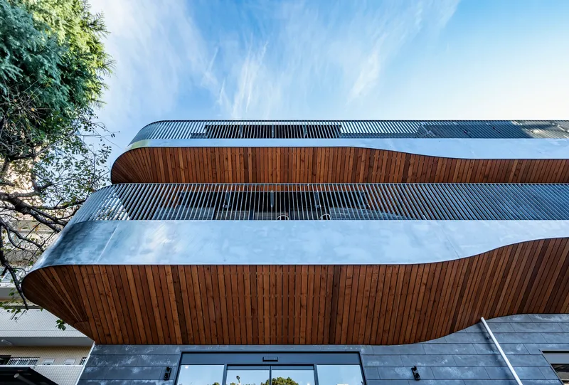 Street view of the curved facade with alternating wood and vertical metal slat bands beneath wispy clouds