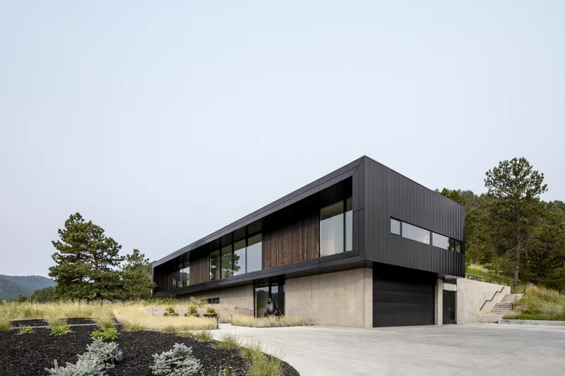 Two-story volume with black corrugated metal upper level and concrete lower level beside a concrete driveway