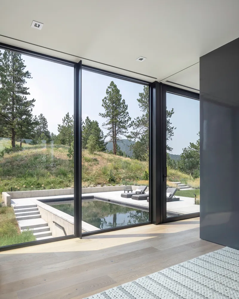 Interior view through black-framed glazing to an exterior pool terrace with pine trees beyond