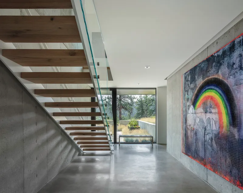 Open tread timber staircase with glass balustrade rises beside a concrete wall displaying a rainbow tapestry