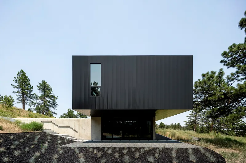 Dark metal clad volume perches above a concrete plinth with open carport cut into the hillside
