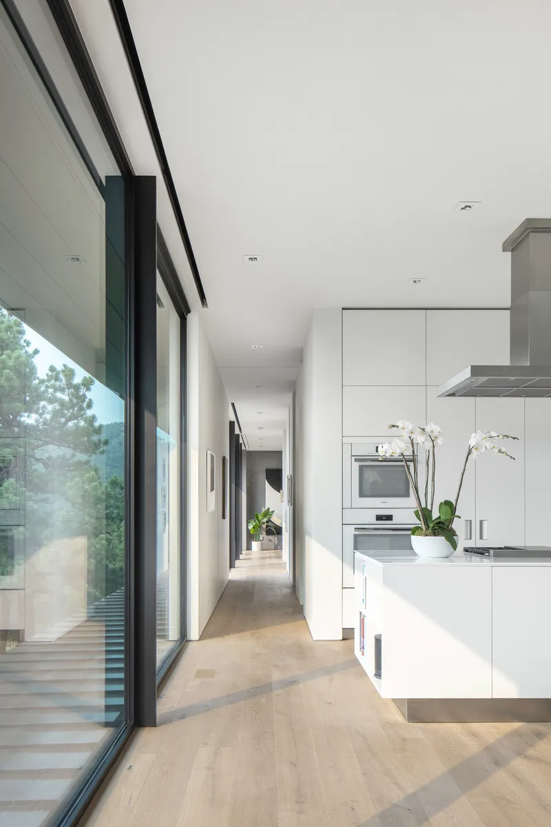 Galley kitchen with white cabinetry along a corridor with continuous glazing overlooking trees