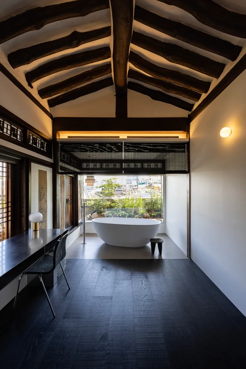 Freestanding bathtub beneath exposed timber rafters with a view to the planted courtyard
