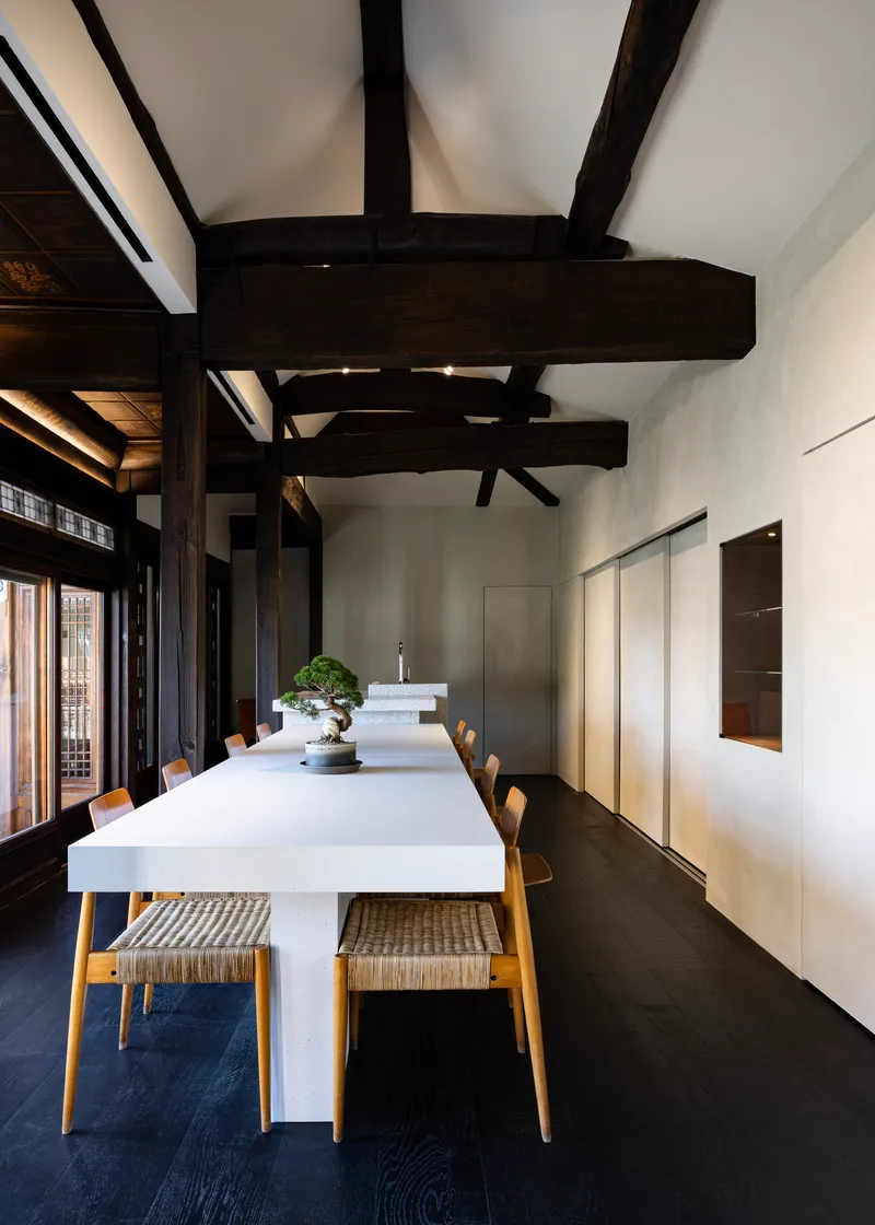 Dining room with exposed dark timber beams above a white table and wooden chairs on dark flooring