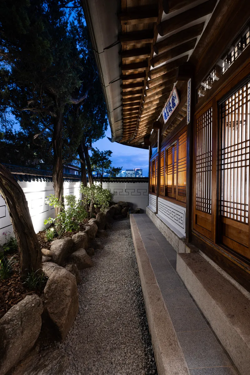 Covered veranda with exposed timber rafters and latticed screen doors overlooking gravel garden at twilight