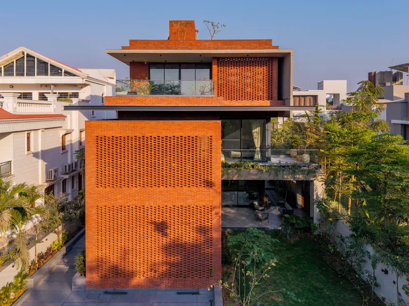 Three-story brick facade with perforated screen walls and rooftop terrace amid neighboring residential structures