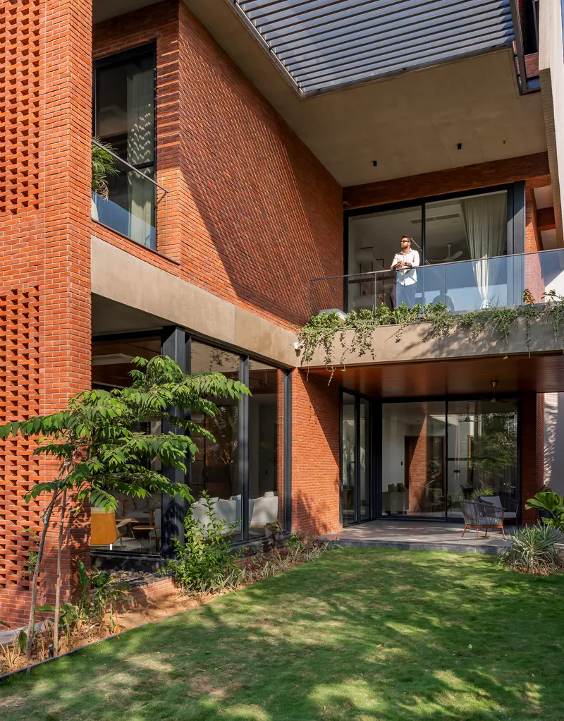 Perforated brick facade with planted balcony where a resident stands overlooking the garden below