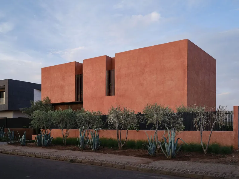 Side elevation of terracotta volumes with vertical screens and front garden of olive trees at dusk