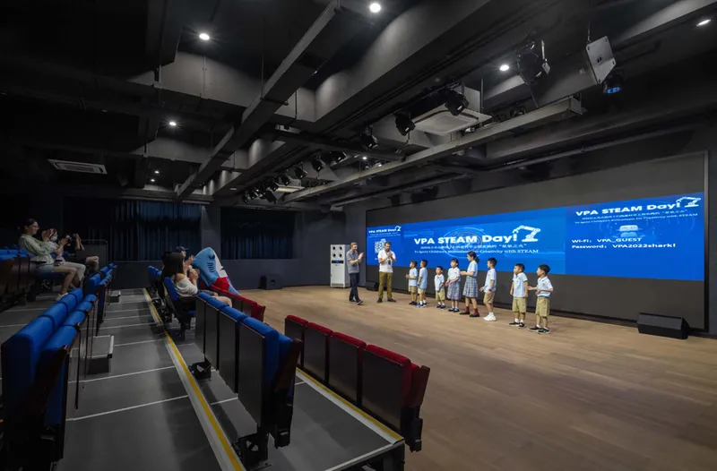 Auditorium with tiered seating and exposed black ceiling facing a blue projection screen during a presentation