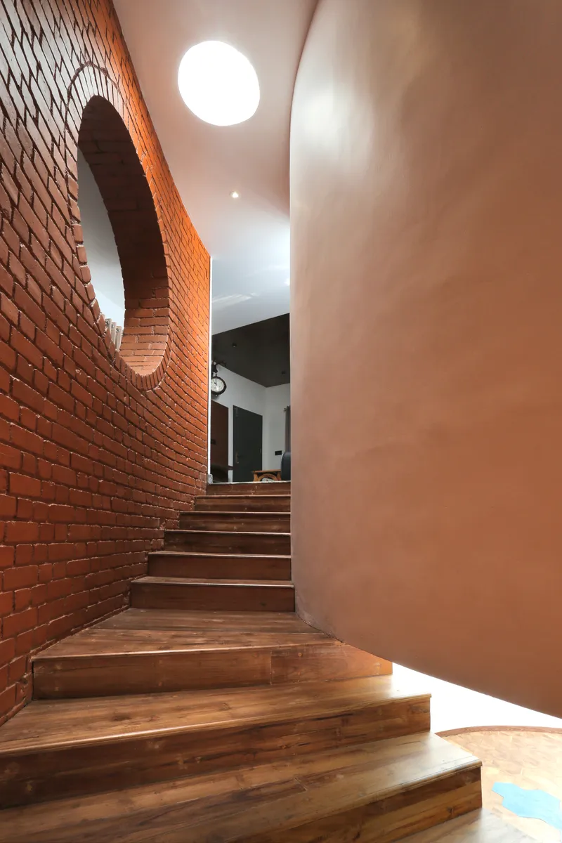Timber staircase ascending between a curving red brick wall and a smooth plastered column lit by ceiling oculus
