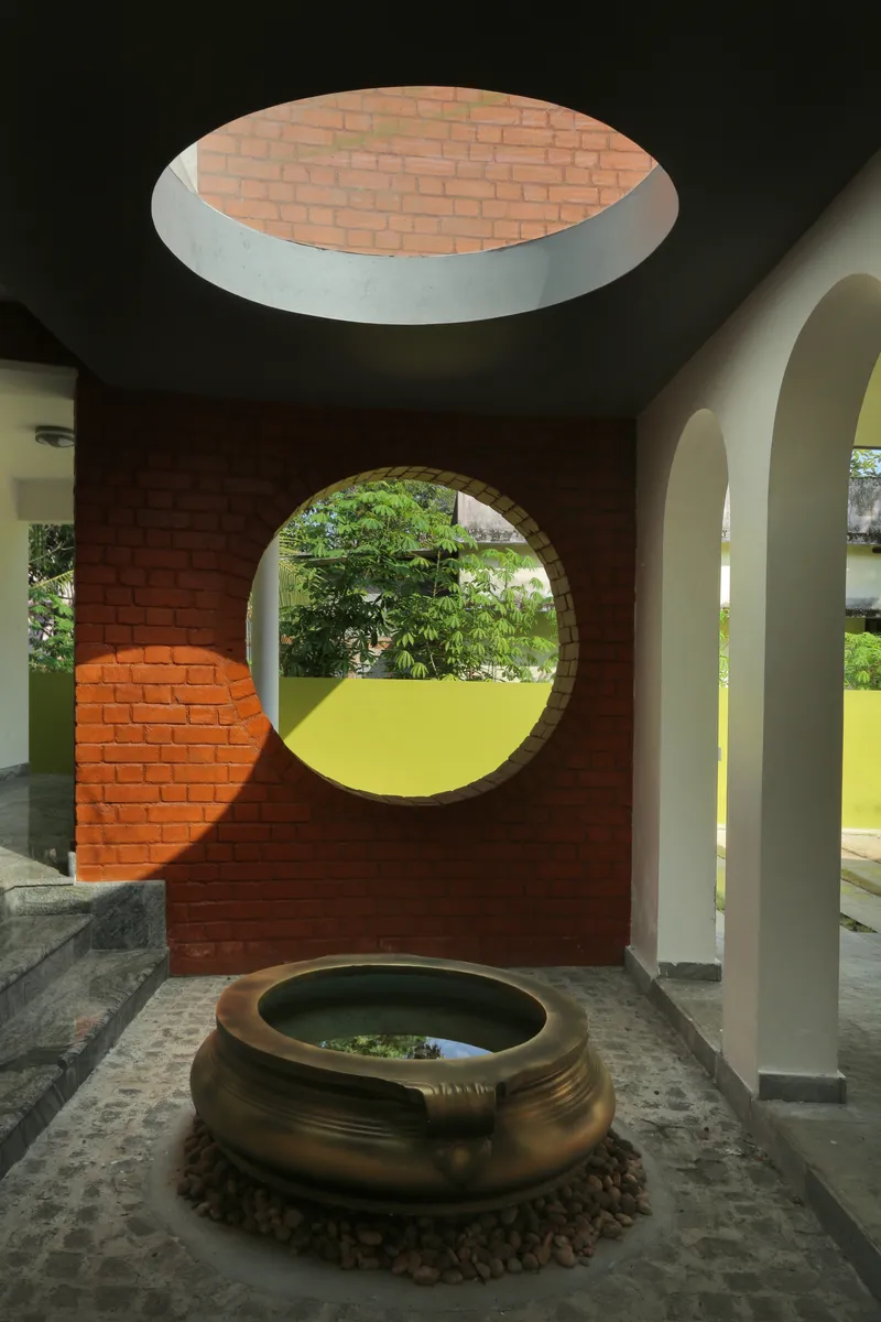 Circular brick oculus in ceiling above a brass water vessel and round portal opening to green landscape