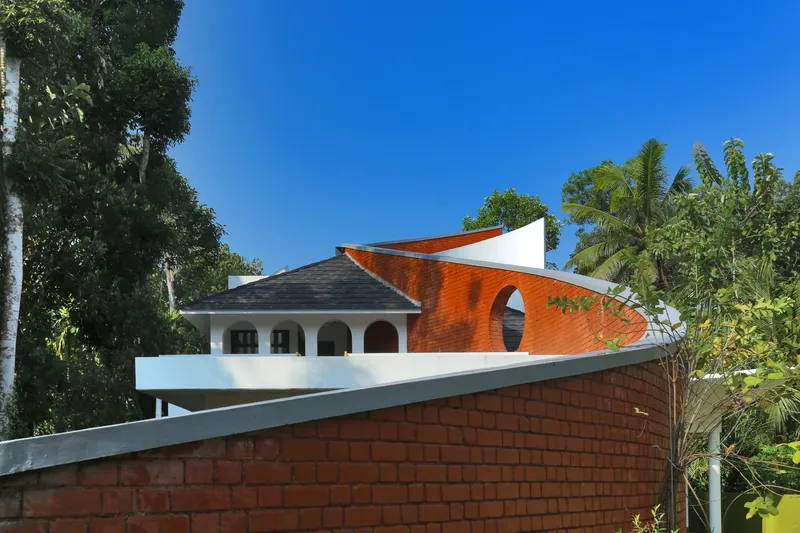 View of red brick boundary wall with the curved white and terracotta roof forms rising beyond palm trees