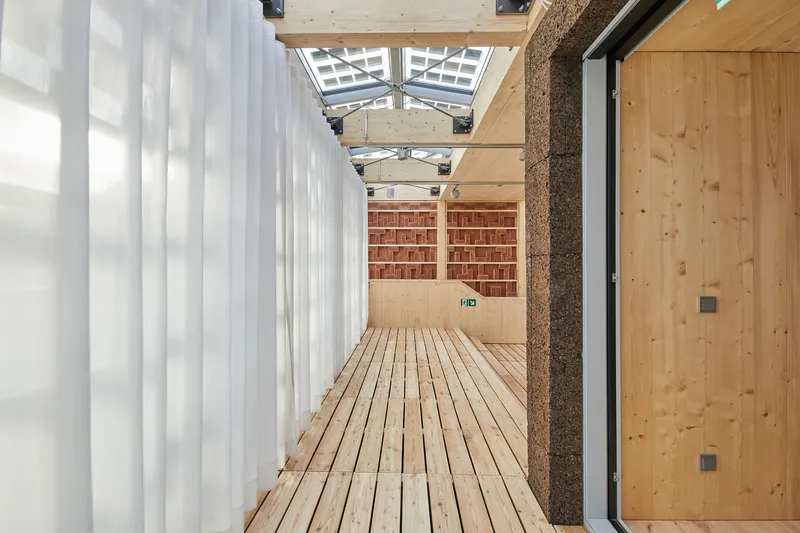 Timber deck corridor with white curtains and cork wall leading toward brick storage
