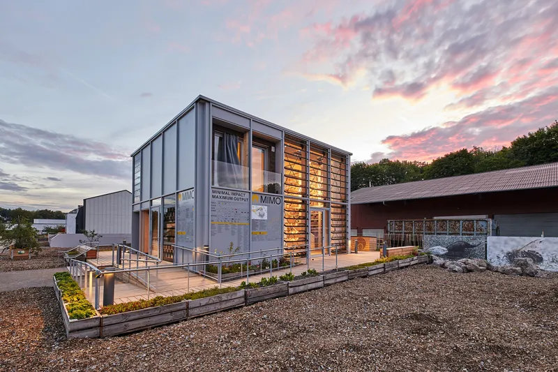 Two-storey corrugated metal structure with timber screen facade and planted beds under a pink sunset sky