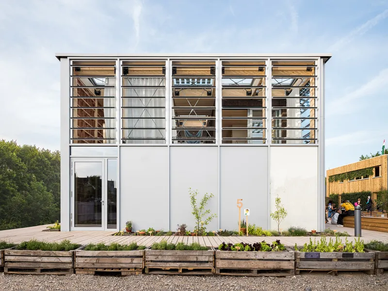 White panel facade with horizontal louvers above glass doors and planted beds with young saplings