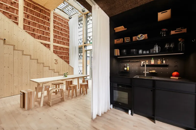 Interior showing exposed brick wall, light wood dining area and black kitchenette separated by white curtain