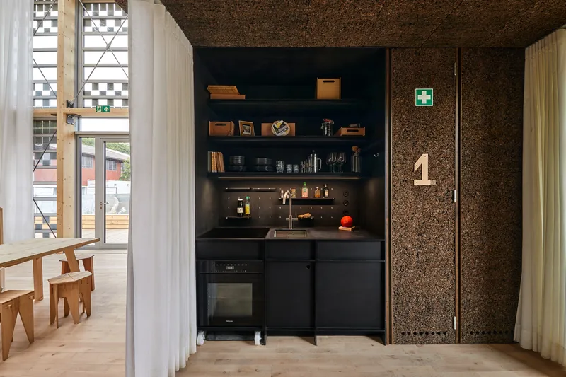Black kitchenette with open shelving next to cork wall panel and white curtain divider