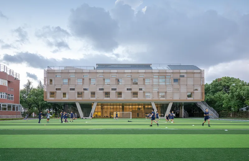 Elevated volume on angled concrete columns above a green sports field with students playing beneath overcast skies