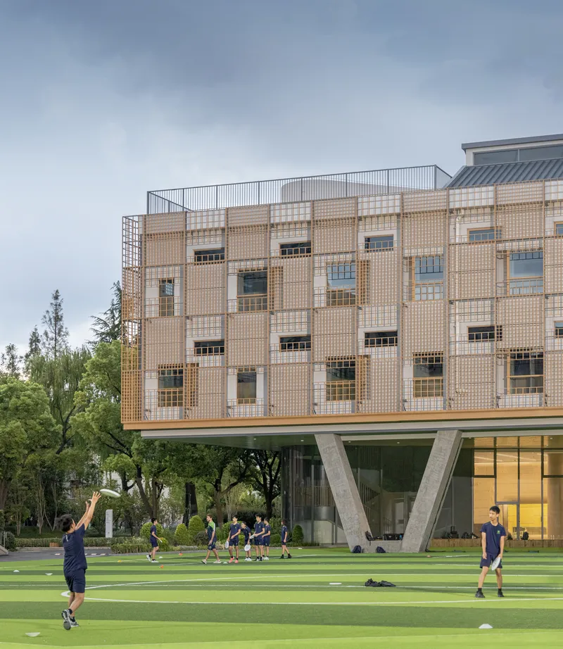 Facade of perforated tile panels and windows suspended over glazed ground level with people on the field below