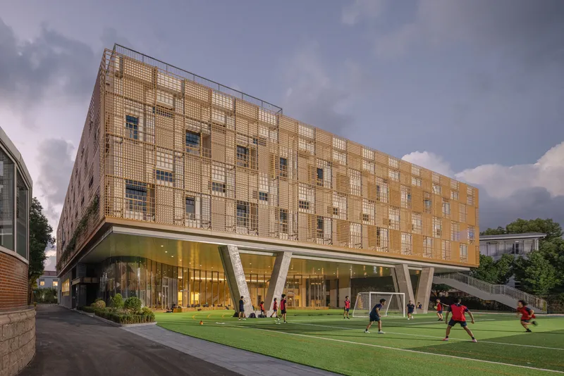 Perforated metal facade above glazed ground level with students on synthetic turf at dusk