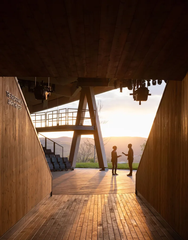 Covered timber deck with angled structural columns framing two silhouetted figures at sunset