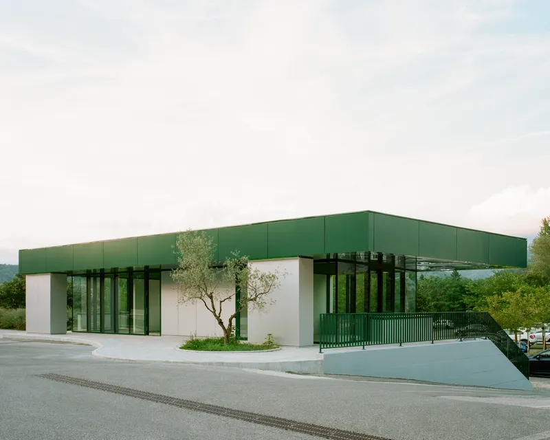 Long view of the green panel roof floating above glass and white volumes beside a roadway
