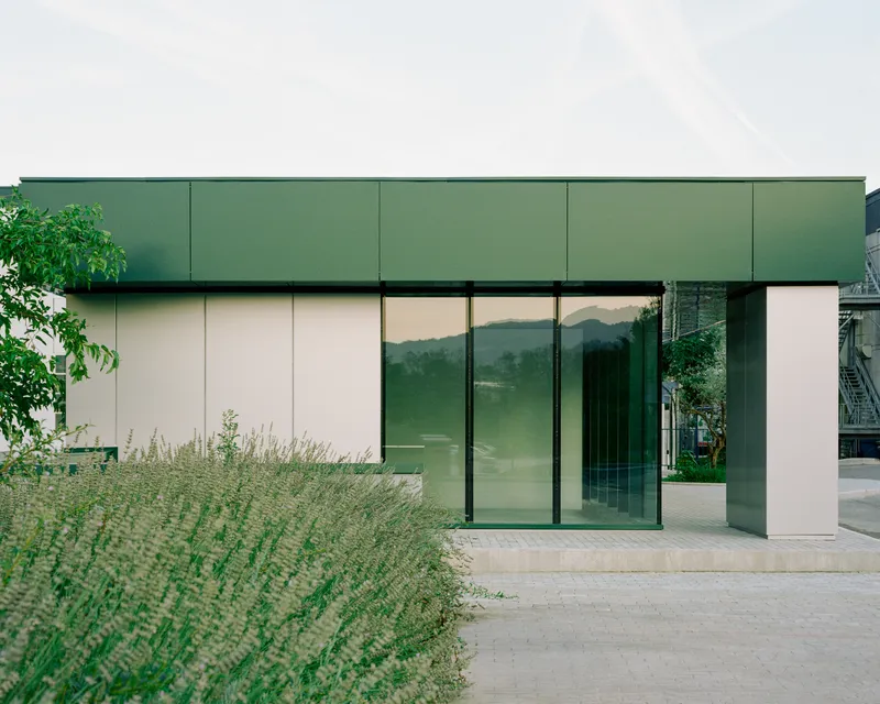 Green metal paneled upper volume above glass entrance doors with planted grasses alongside paved terrace