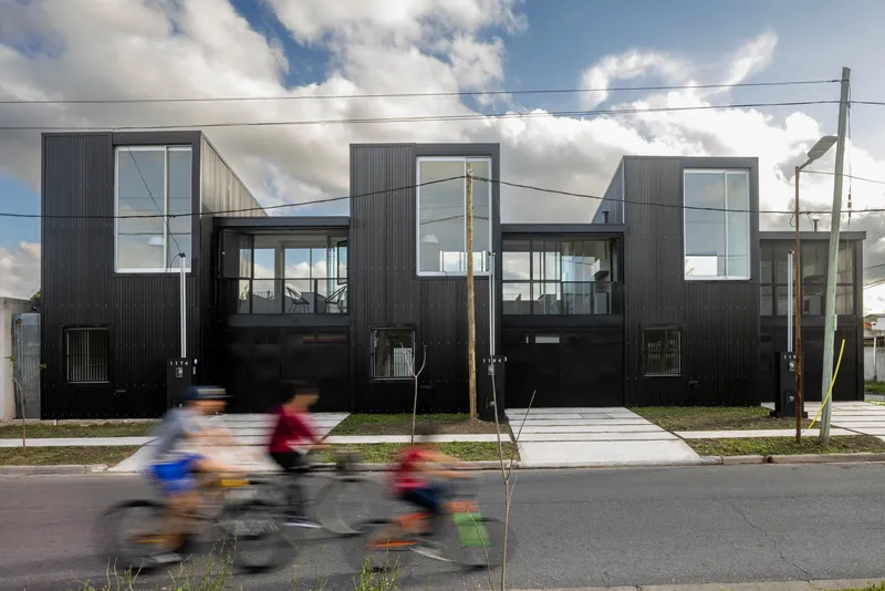 Street view of three black corrugated metal townhouses with cyclists passing in front on a cloudy day