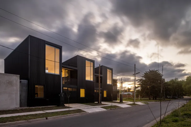 Facade of black corrugated townhouses with glowing interiors visible through tall windows at twilight