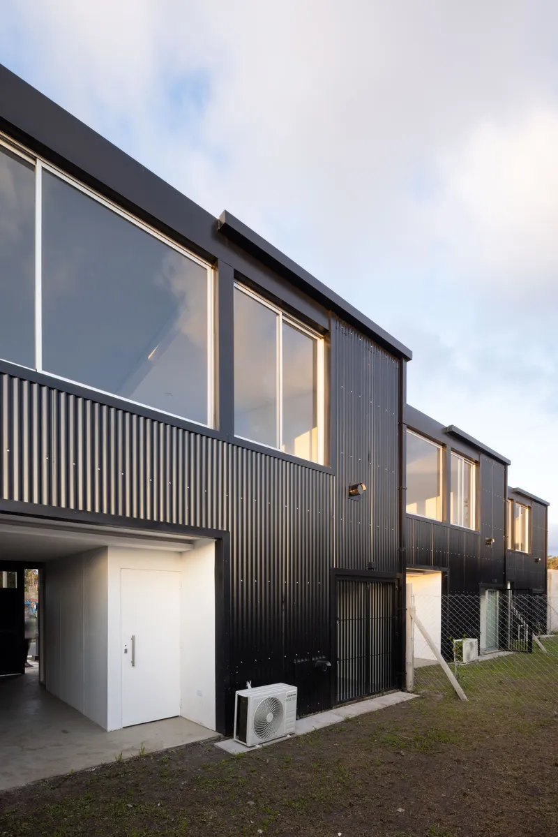 Row of black corrugated metal facades with upper glazing and ground-level carports under overcast sky