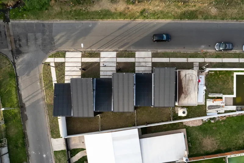 Drone view of the linear townhouse roofline with dark corrugated metal panels and courtyard voids beside the street