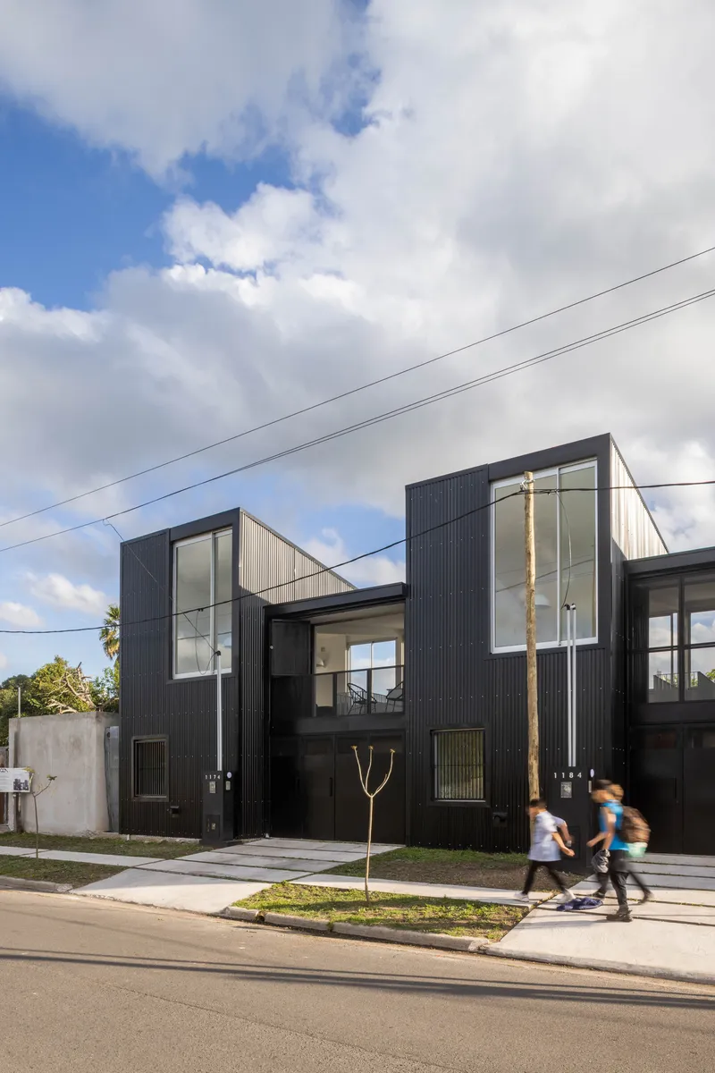 Street view of the black corrugated metal facade with glazed volumes and two people cycling past on the sidewalk