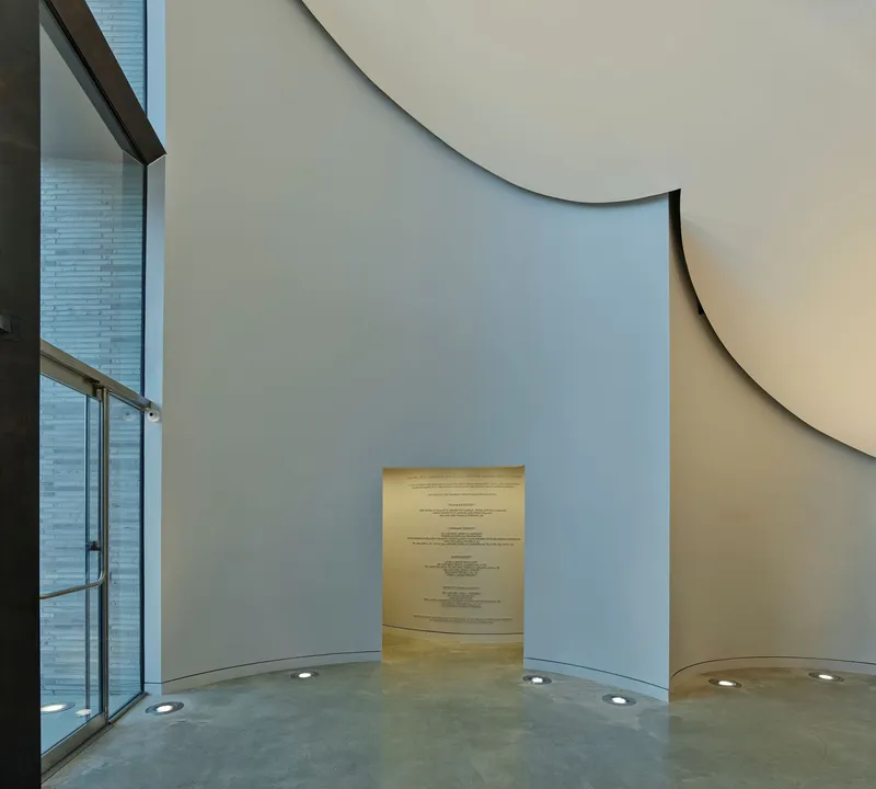 Curved interior wall with narrow illuminated doorway and floor-recessed uplights beside glazed facade