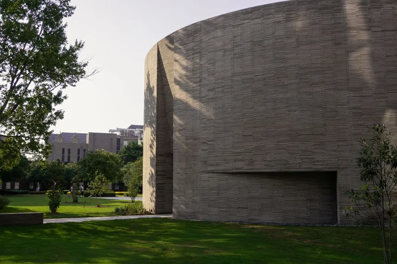 Curved brick chapel volume rising above a green lawn with trees casting afternoon shadows