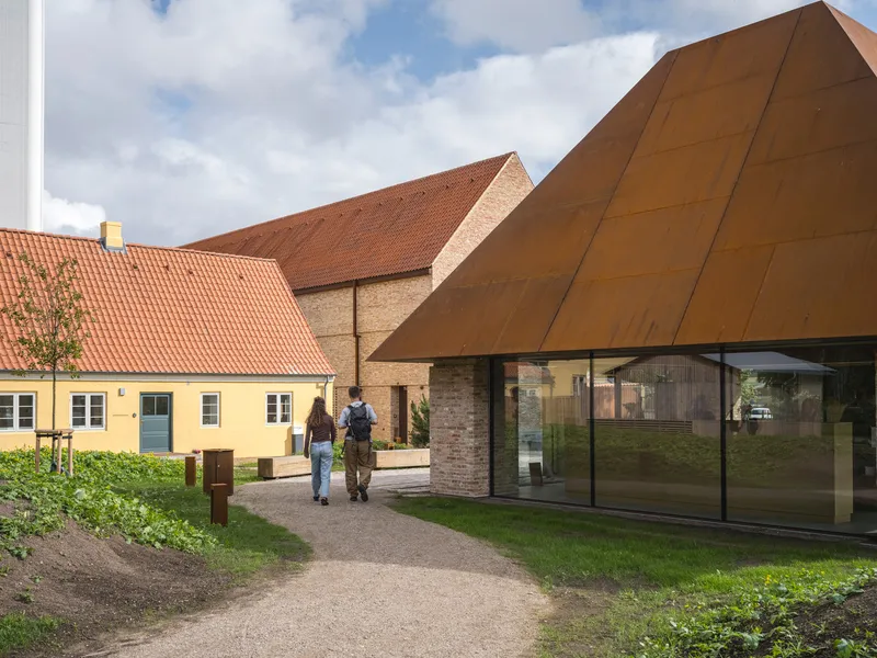Two visitors walking a gravel path past the angled Corten steel facade with large glazed entry