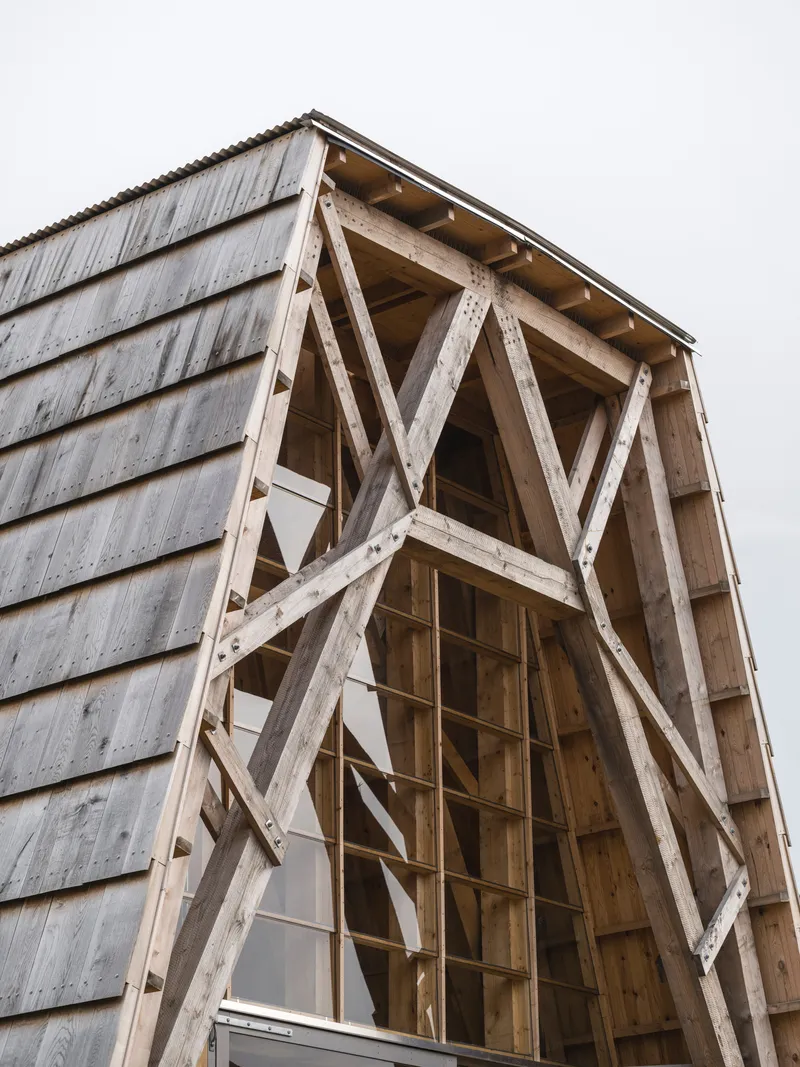 Exposed timber diagonal bracing within a gabled facade opening clad in weathered horizontal siding