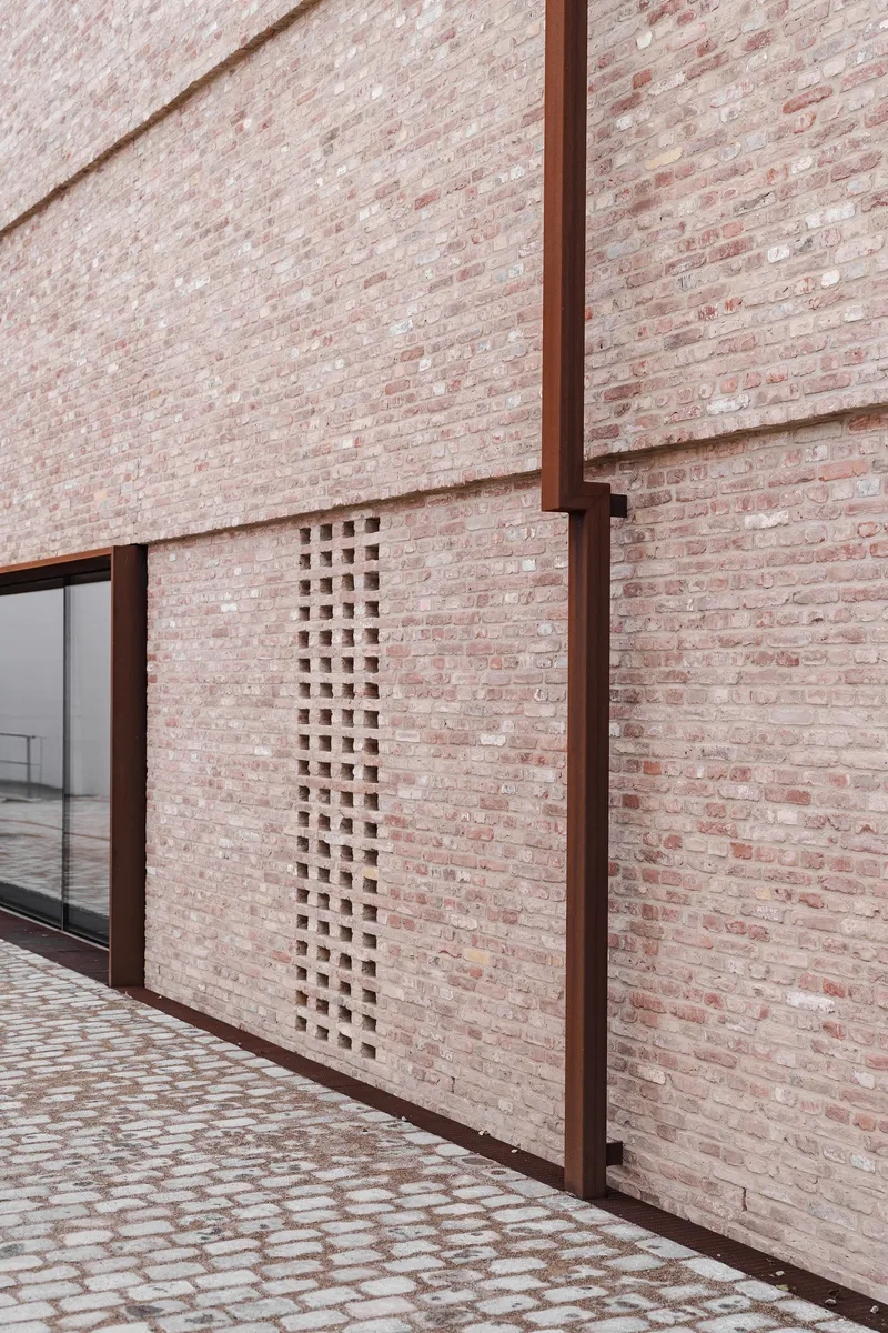 Brick wall with vertical perforated screen and steel frame along cobblestone courtyard