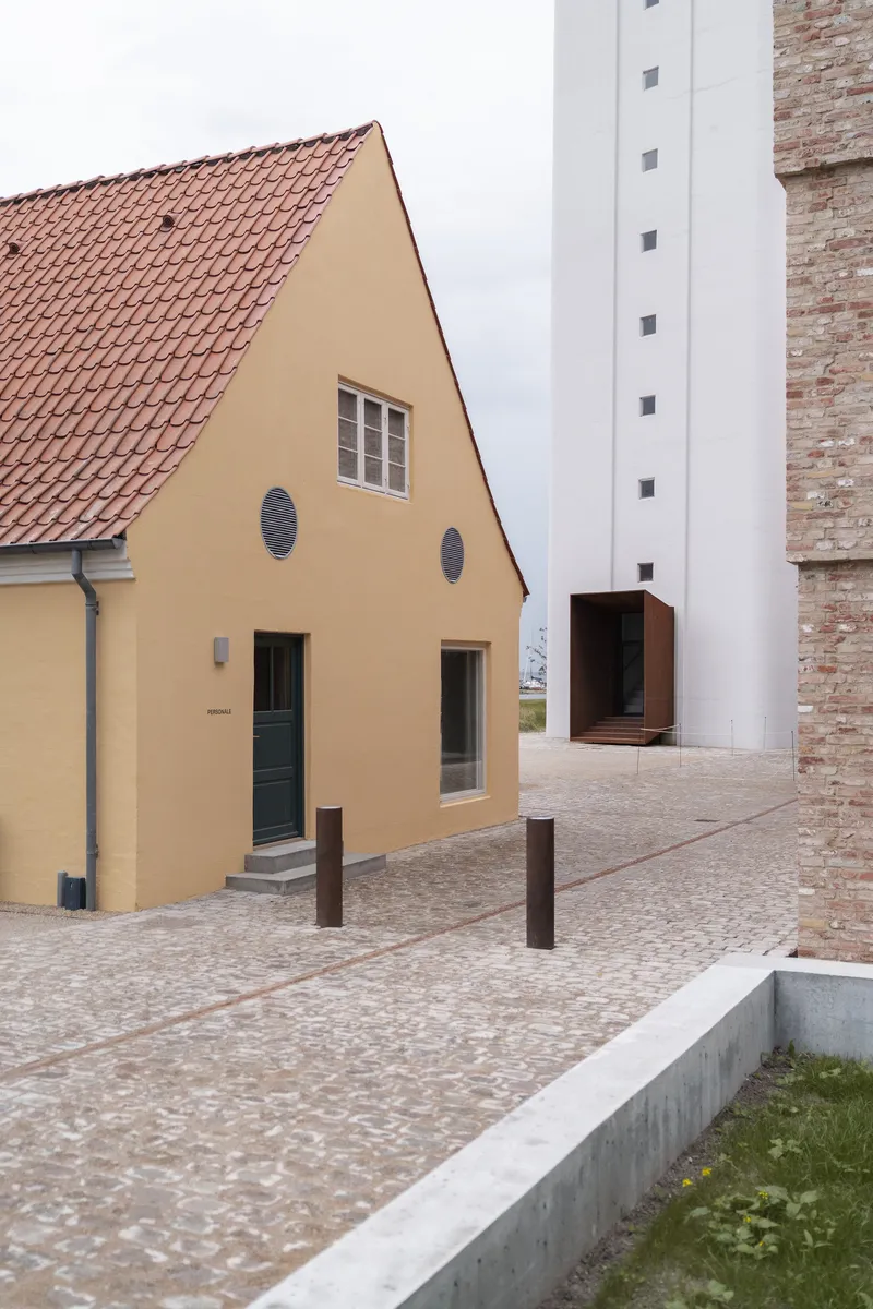 Cobblestone courtyard with yellow gabled house, white tower, and bollards under an overcast sky