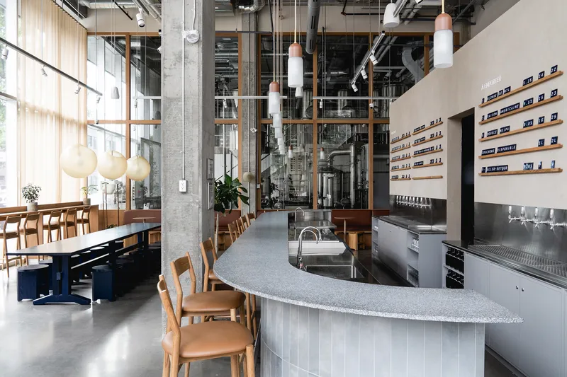 Open brewery interior with curved terrazzo bar, exposed concrete columns, timber-framed glazing and pendant lights