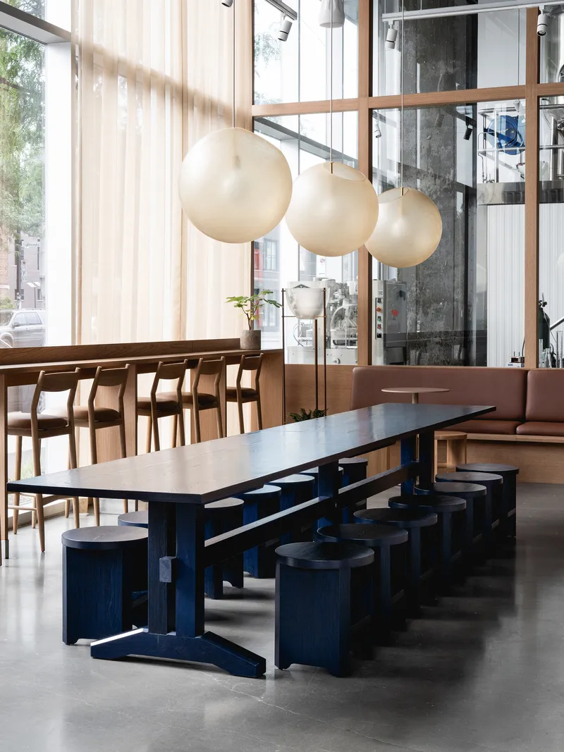 Communal table in navy-painted timber with cylindrical stools beneath clustered globe pendants and sheer curtains