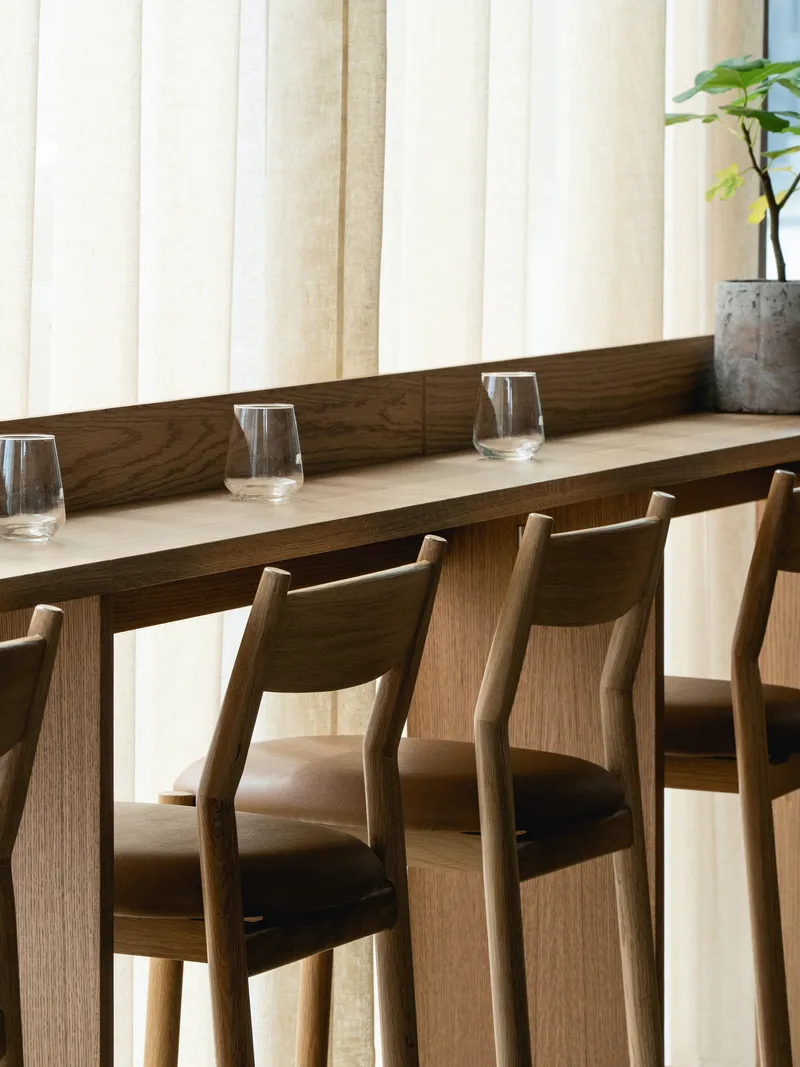 Oak dining table with angled-back chairs and glassware against sheer linen curtains in soft daylight