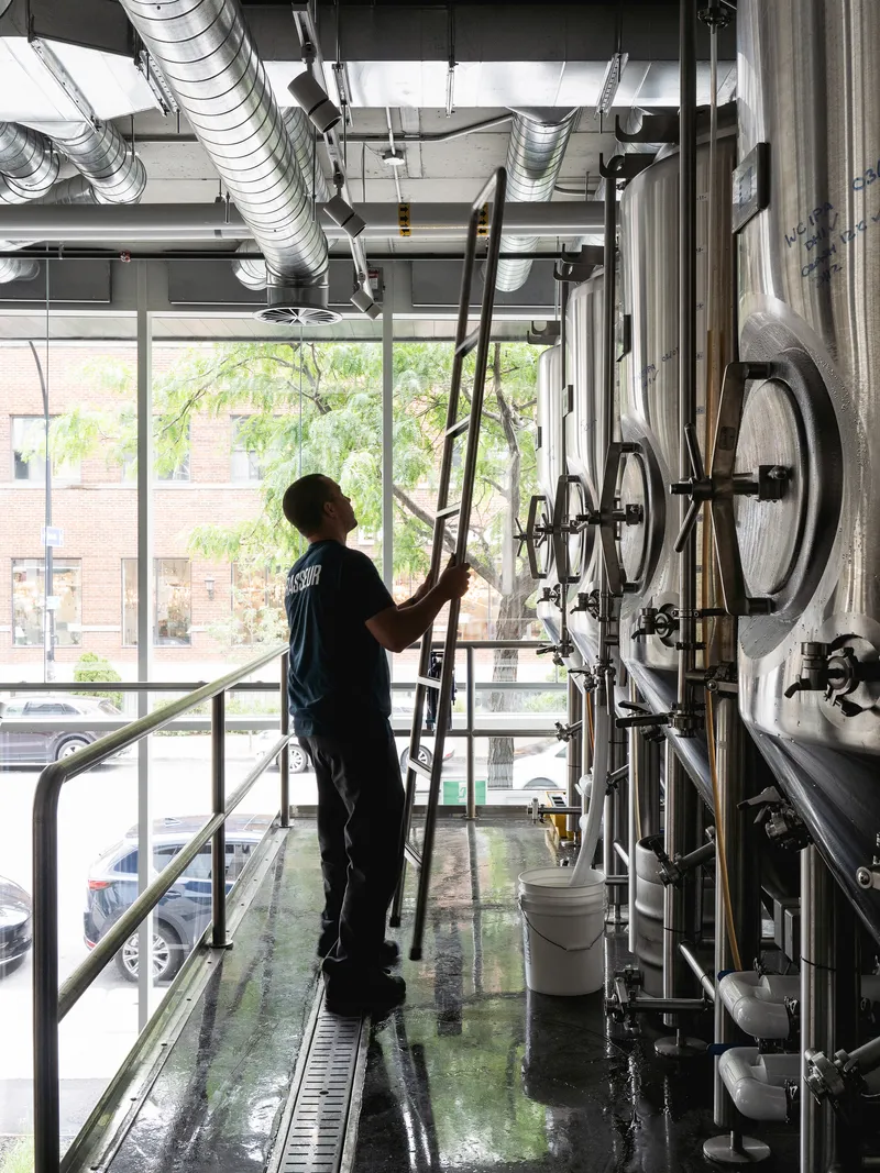 Worker on a ladder among exposed ductwork and stainless steel brewing tanks with trees visible through the windows