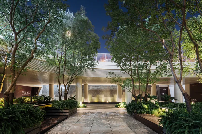 Evening view of illuminated entry pavilion with stone walls and uplighting beneath established trees at dusk