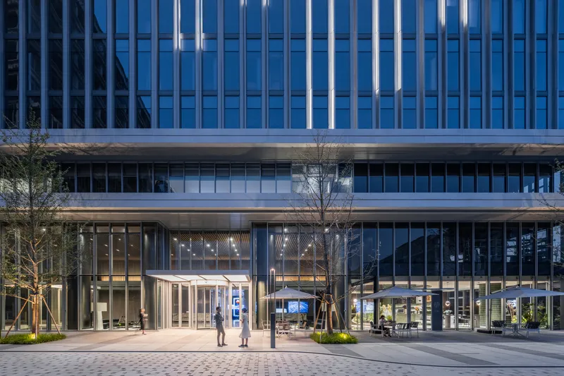 Ground level plaza with glass entrance pavilion, paired figures walking, and young trees at dusk