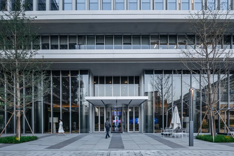 Entrance canopy with glass walls flanked by young trees and a person walking in early evening
