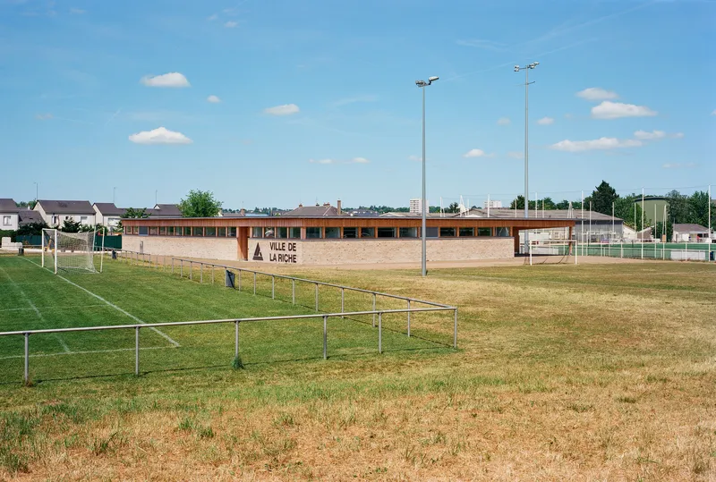 View across sports pitches showing the low horizontal volume with timber and brick facade under summer sky